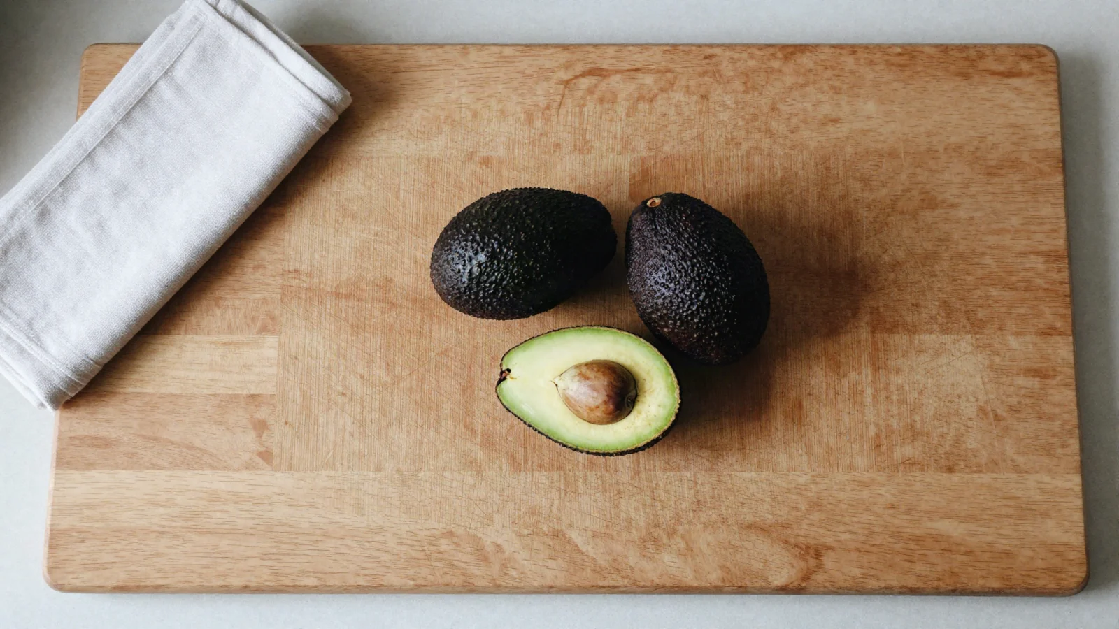 Two whole avocados and a halved avocado on a wooden cutting board.