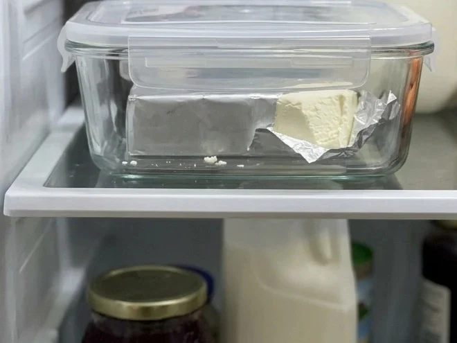How to store cream cheese in the fridge: a partly used block in its folded foil inside a lidded glass container on a shelf.