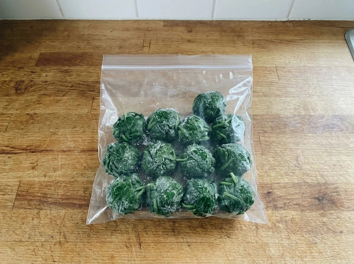 Frosted balls of cooked spinach in a flat zip-top freezer bag on a counter.