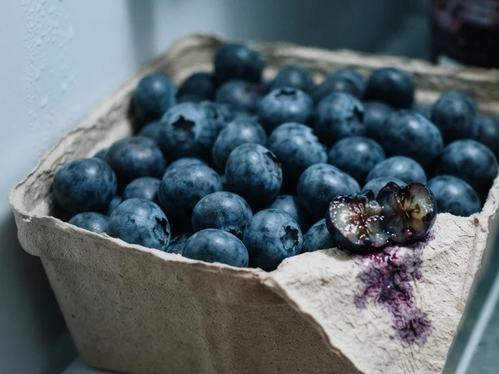 A split blueberry at the front of a punnet with a small purple stain spreading beneath it.