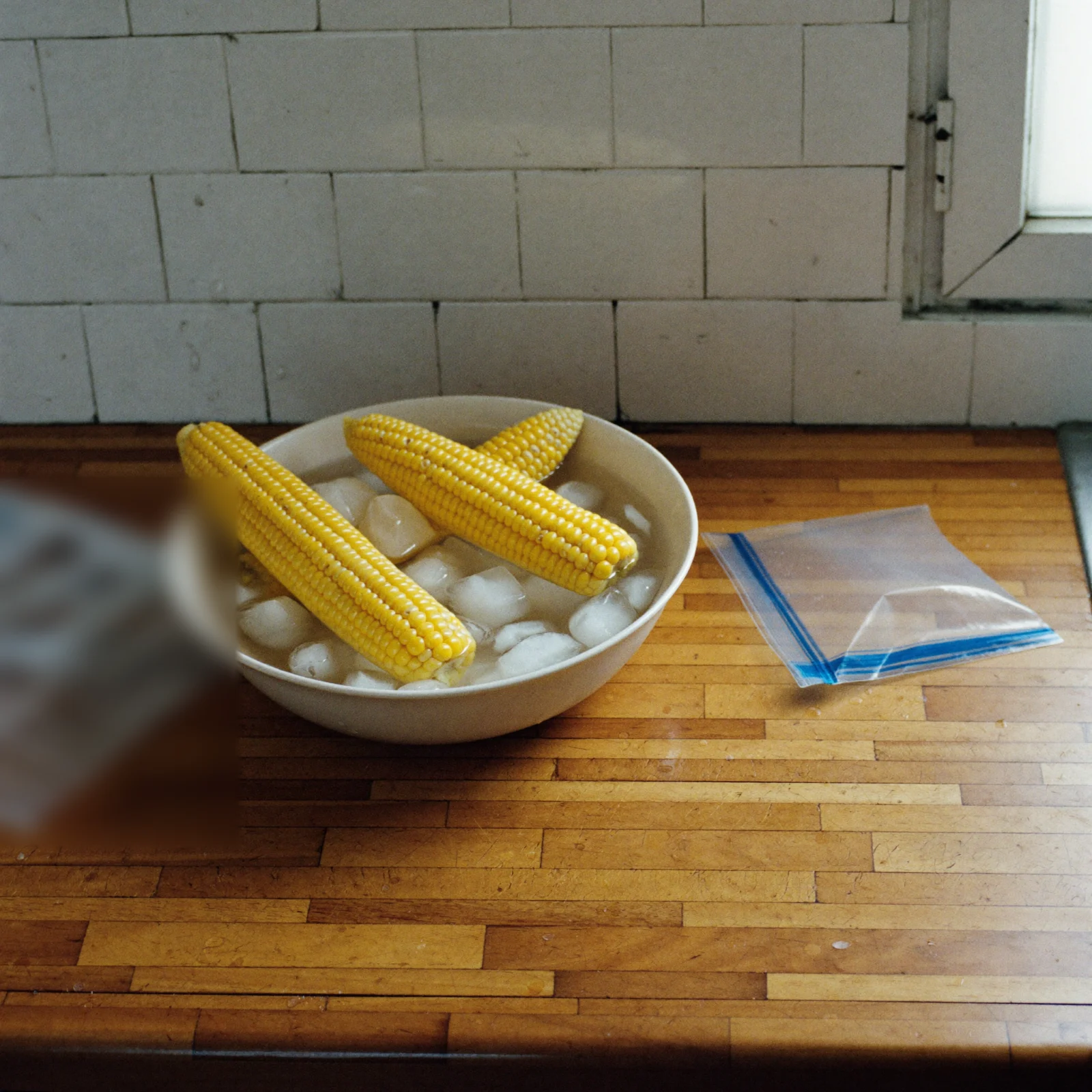 Blanched ears of corn cooling in a bowl of iced water beside a freezer bag
