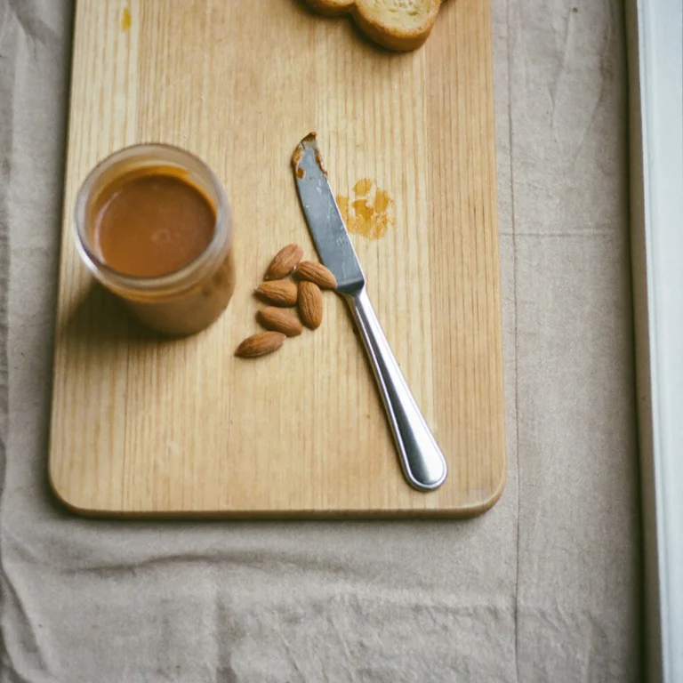 An open jar of almond butter on a beech-wood board with whole almonds, a butter knife and a slice of toast