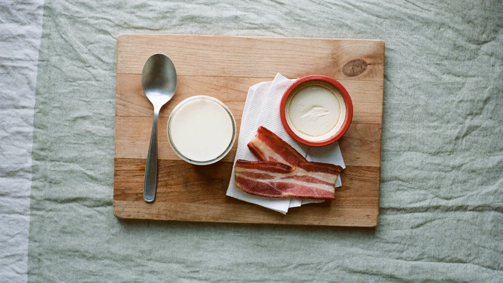 How to Store Bacon Grease - a glass jar of creamy white bacon grease beside crisp bacon strips on a beech-wood board