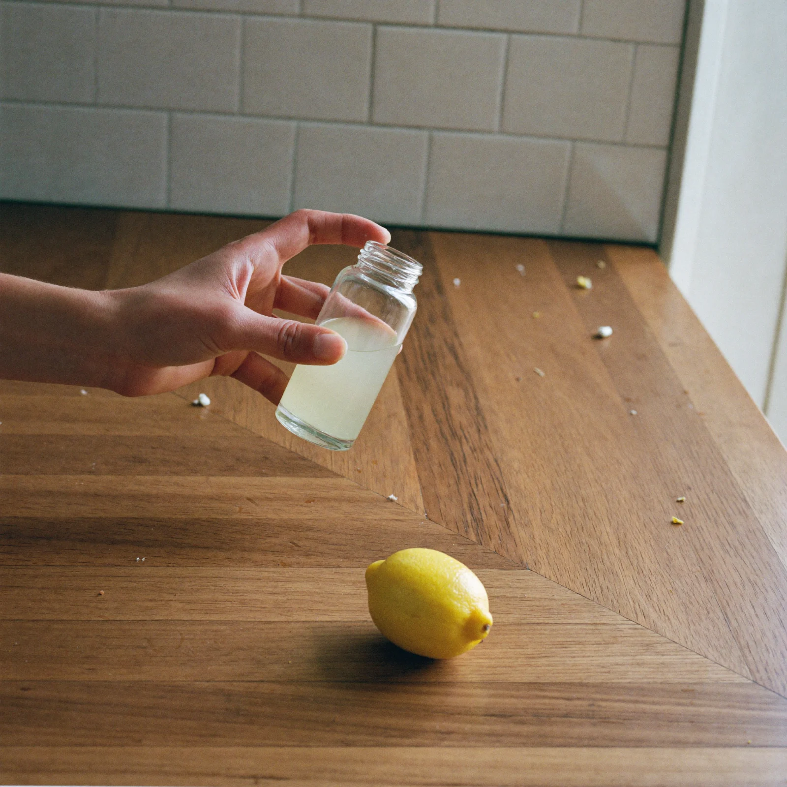A hand holding a small open jar of very pale lemon extract above the counter, with a fresh lemon below