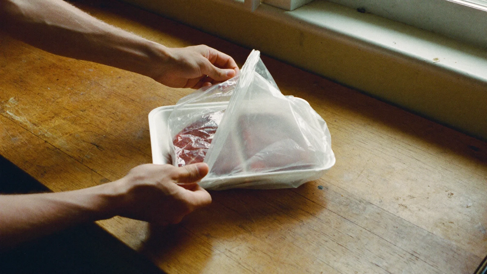 Hands sliding a tray of raw steak into a clear plastic bag to catch leaks