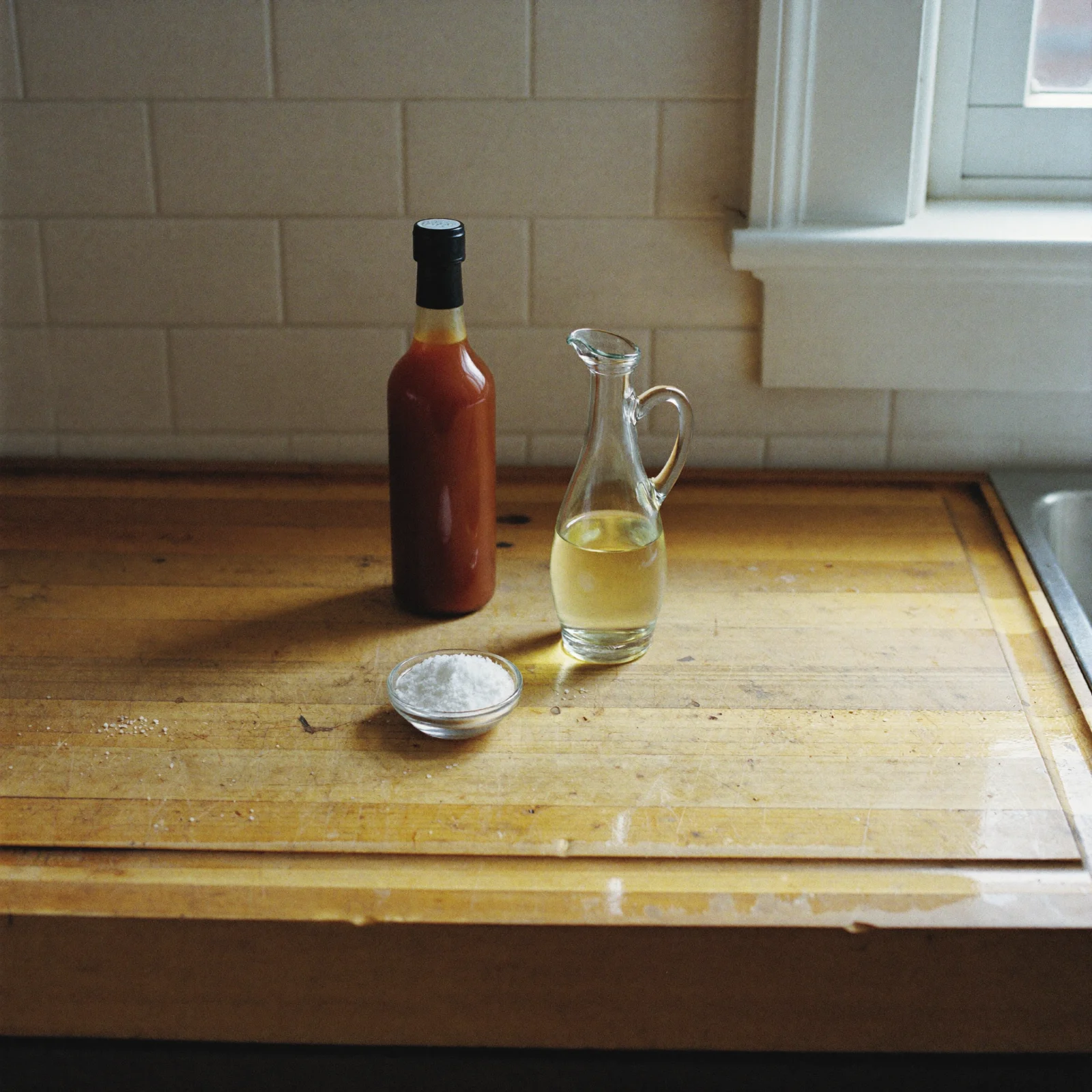 A bottle of brown sauce beside a jug of vinegar and a dish of salt on a kitchen counter